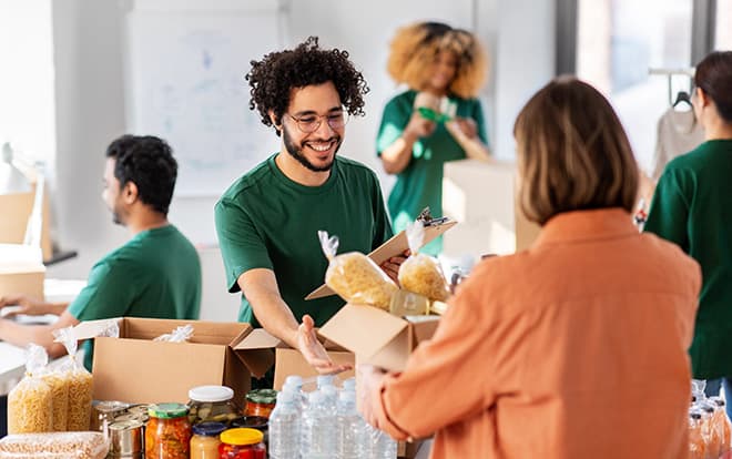 charity, donation and volunteering concept - happy smiling male volunteer with clipboard and woman taking box of food at distribution or refugee assistance center