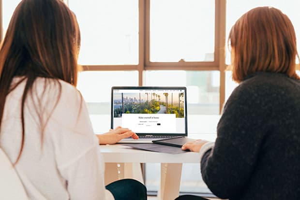 The back of 2 business women sitting at a table looking at a laptop, by a window.