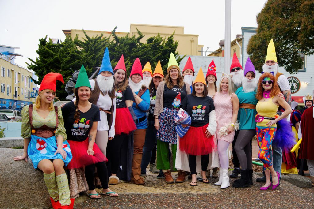 A group of people dressed up as elves. They are smiling and posing for the photo in front of a shopping center on a rainy day.